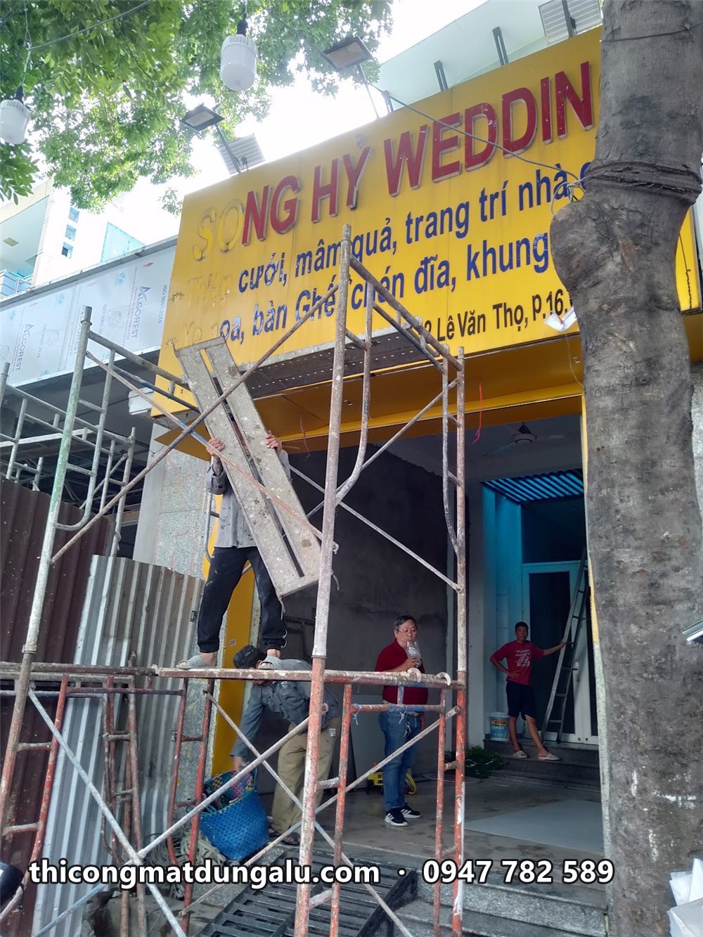 Thợ trên giàn giáo tháo biển cũ Song Hỷ Wedding nền vàng chữ xanh