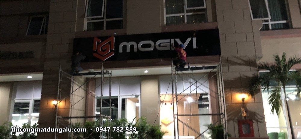 Bảng hiệu Mogivi nền đen logo đỏ trên lối vào tòa nhà lúc chiều tối