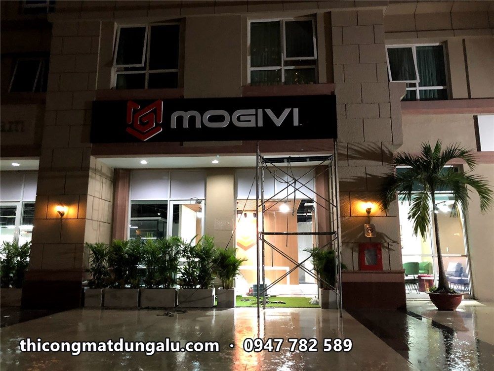Tòa nhà với bảng hiệu Mogivi đen, cây xanh trước sảnh, giàn giáo còn ở bên phải
