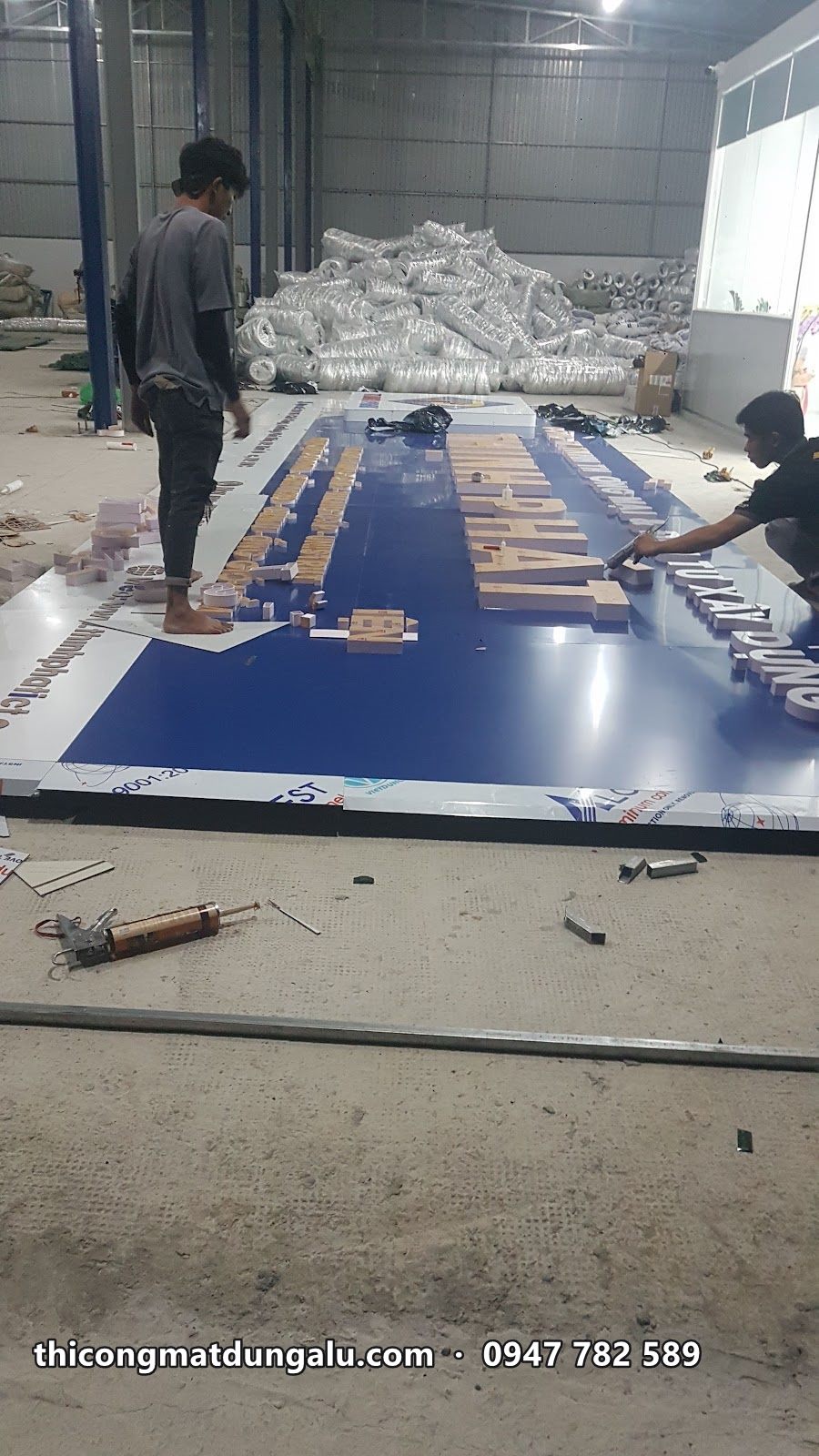 Hai thợ gắn chữ lên mặt alu xanh dài trải dọc sàn xưởng