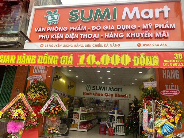 Bảng hiệu SUMI Mart nền cam chữ nổi xanh trắng, dòng danh mục hàng và băng rôn gian hàng đồng giá