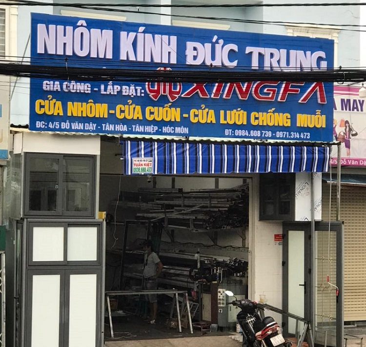 Bảng hiệu cửa hàng nhôm kính nền xanh với logo hãng nhôm và các dòng sản phẩm, cửa mẫu bên dưới