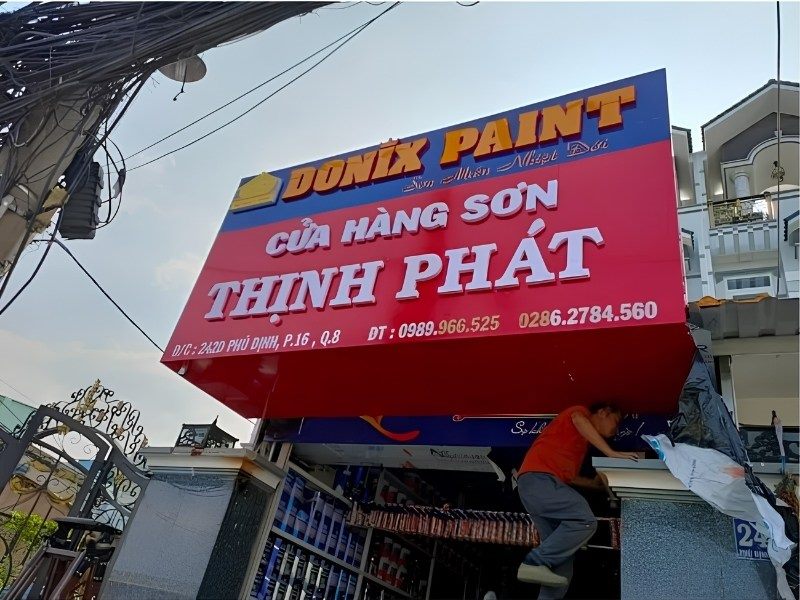 Thợ đang lắp bảng hiệu cửa hàng sơn Thịnh Phát nền đỏ, chữ nổi trắng