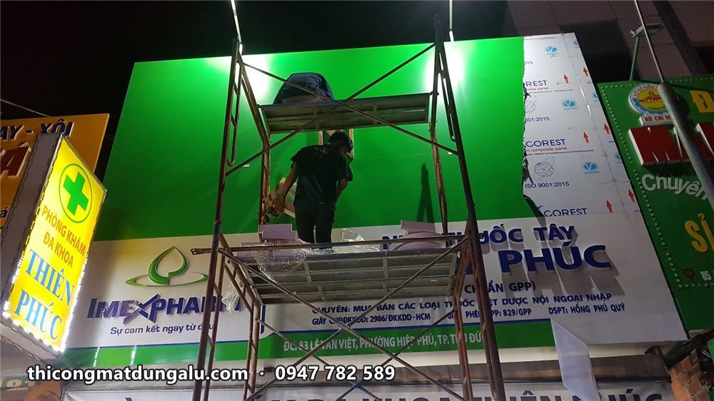 Thợ trên giàn giáo lắp chữ nổi lên mặt tiền alu xanh của phòng khám vào buổi tối