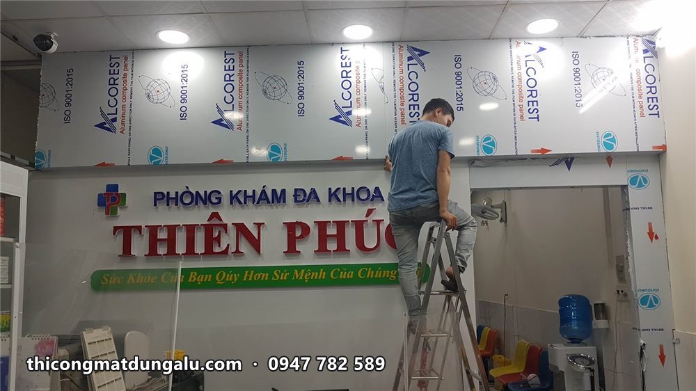 Thợ đứng trên thang gắn chữ nổi tên phòng khám lên tường ốp alu trong sảnh