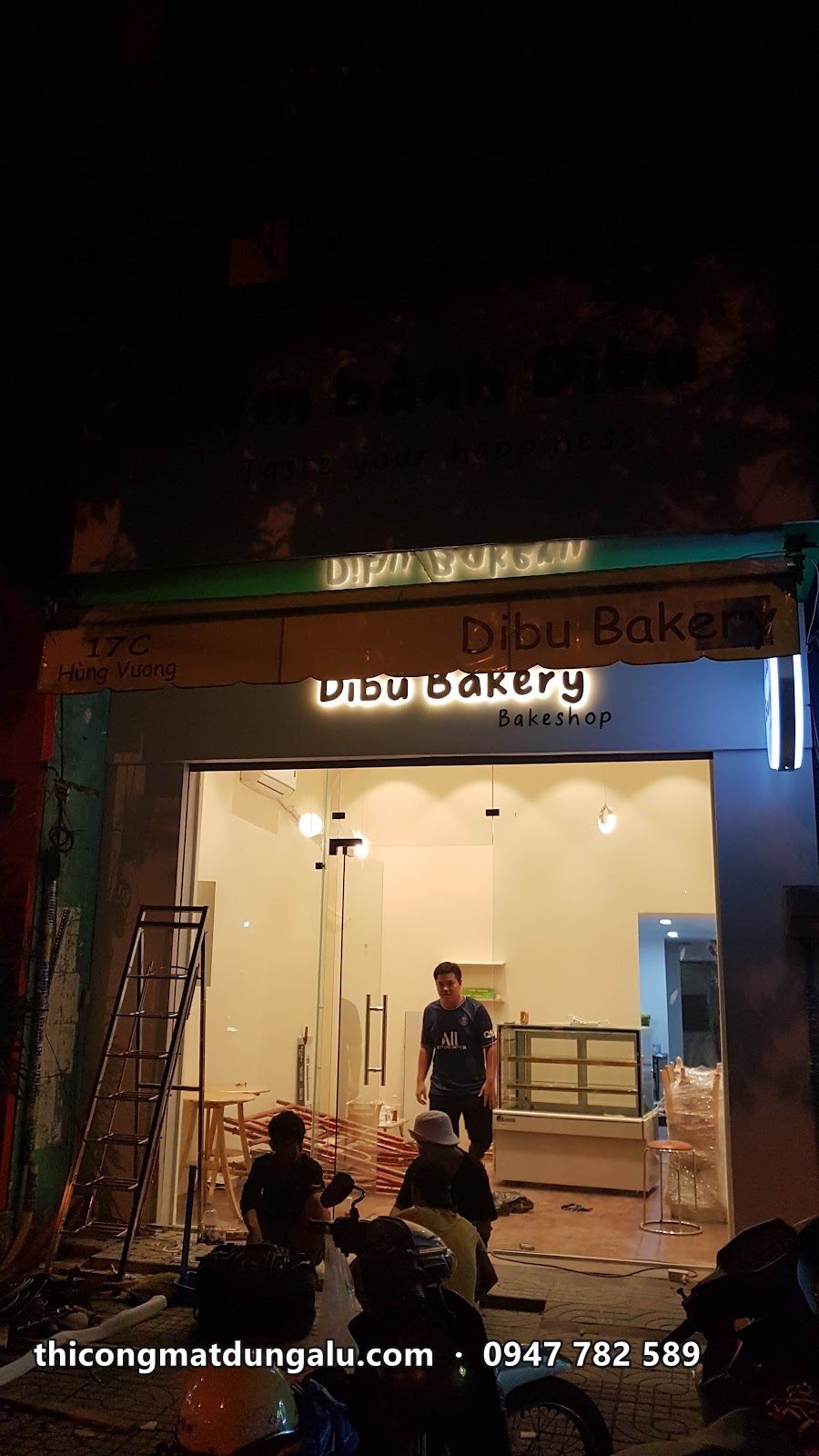 Thợ lắp bảng hiệu Dibu Bakery ban đêm, hộp đèn tròn treo bên phải