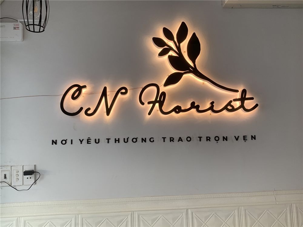 Logo shop hoa chữ viết tay đen hắt sáng vàng cùng cành lá trên tường trắng