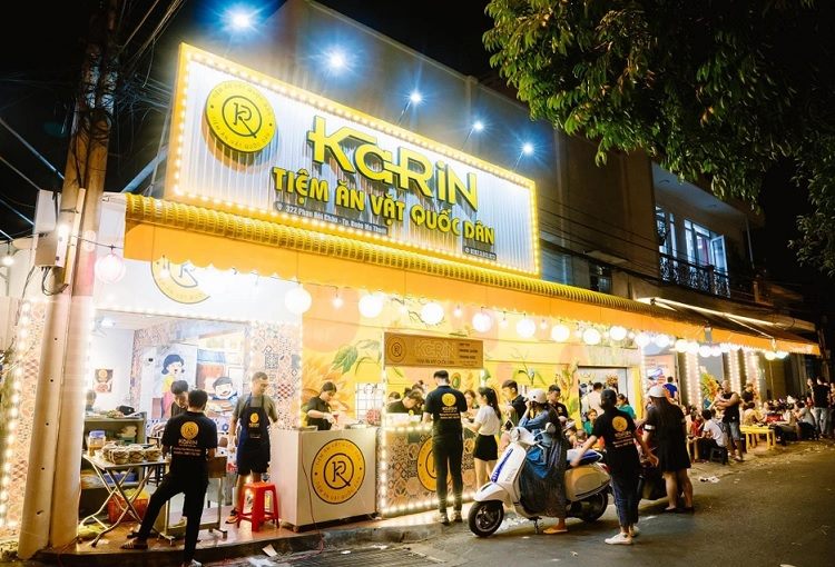Tiệm ăn vặt ban đêm nền vàng, viền bóng đèn sáng, khách đông trước cửa