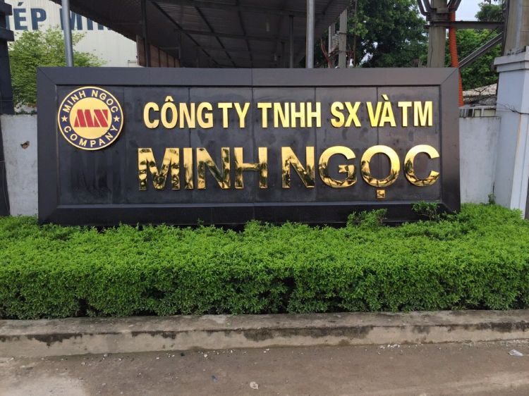 Bảng tên nhà máy nền đen chữ inox vàng và logo, có viền cây xanh phía dưới