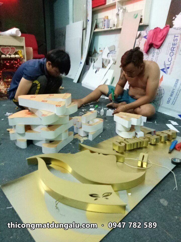 Hai thợ ngồi trên sàn gia công chữ inox vàng và logo cong