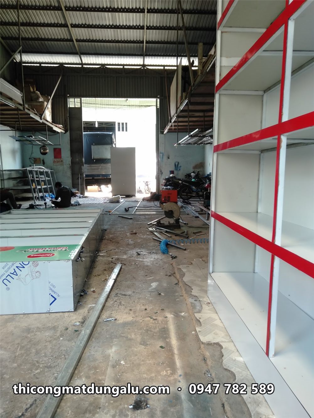 Kệ trưng bày viền đỏ đứng dọc tường xưởng, dãy bàn kệ phía sau