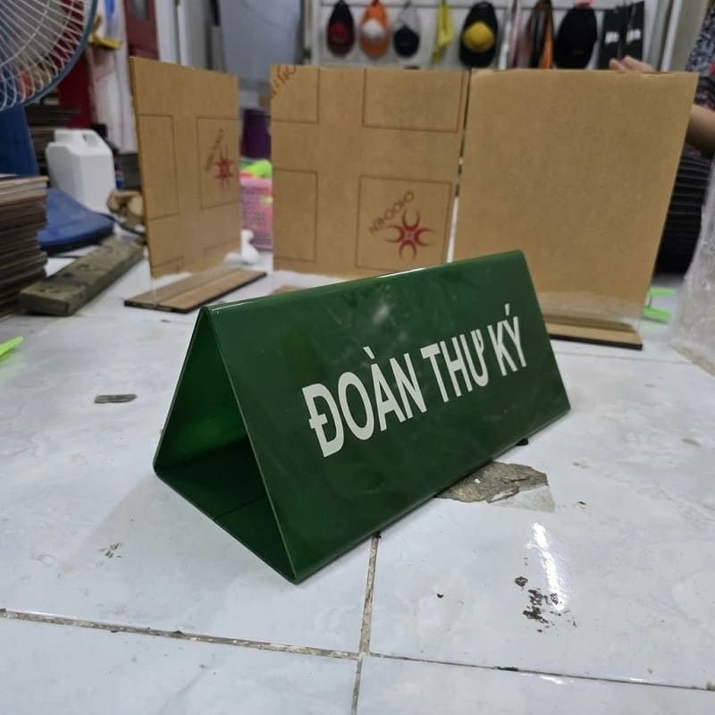 Biển mica chữ A xanh lá đặt trên sàn xưởng, phía sau là tấm mica thô