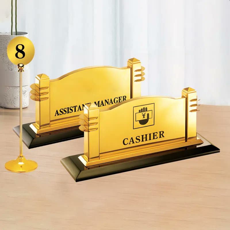 Hai biển chức danh mạ vàng Cashier và Assistant Manager đặt trên bàn gỗ
