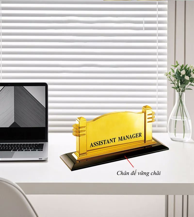 Biển chức danh mạ vàng đặt trên bàn làm việc cạnh máy tính, chú thích chân đế vững chãi