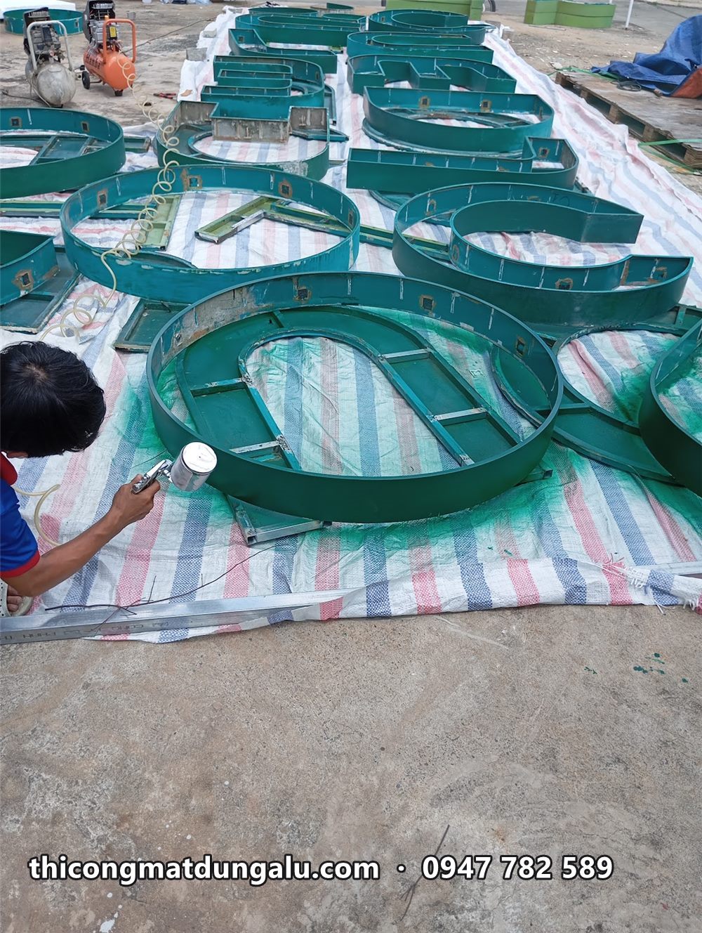 Thợ phun sơn xanh cho hàng loạt khung chữ nổi cỡ lớn trải trên bạt