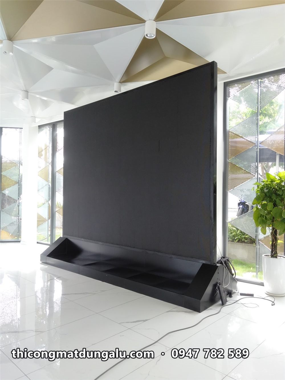 Màn hình LED đen đặt trên chân đế giữa sảnh tòa nhà có trần tam giác, lúc chưa bật