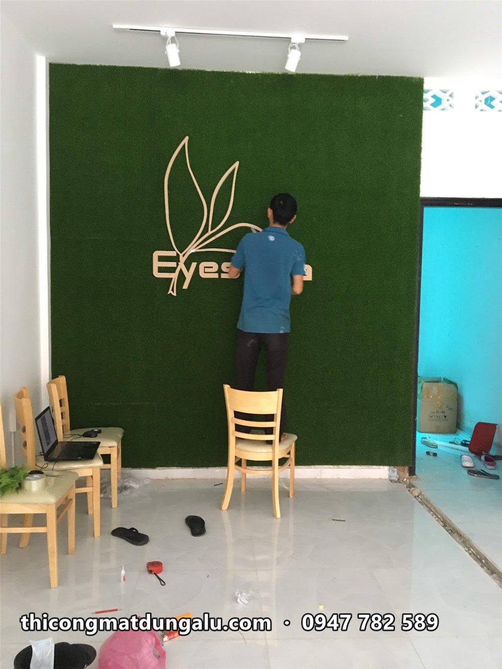 Thợ đứng trên ghế gắn logo Eyes Tea lên tường cỏ nhân tạo