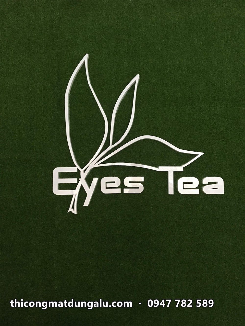 Logo Eyes Tea nét lá trà trắng trên nền tường cỏ xanh