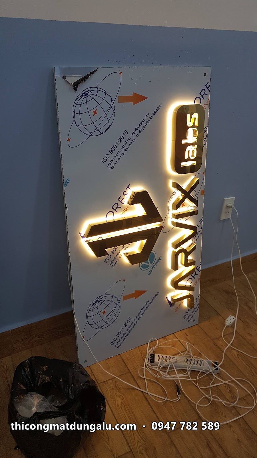 Tấm nền gắn logo inox vàng Jarvix Labs đang thử đèn hắt sáng, dựng tạm trong phòng