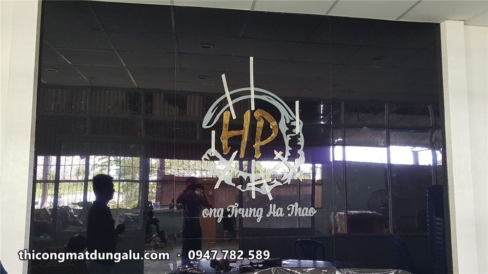 Logo inox vàng HP và dòng chữ tên công ty gắn trên vách kính đen văn phòng, đang hoàn thiện