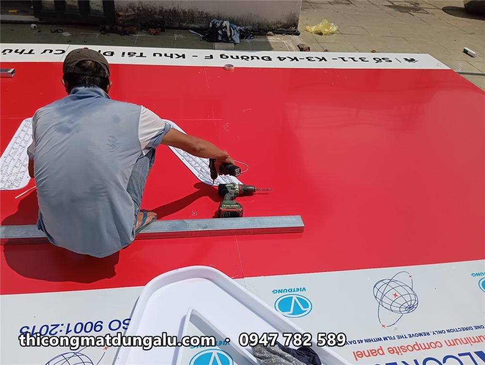 Thợ ngồi trên mặt alu đỏ dùng máy khoan bắt vít thanh nẹp