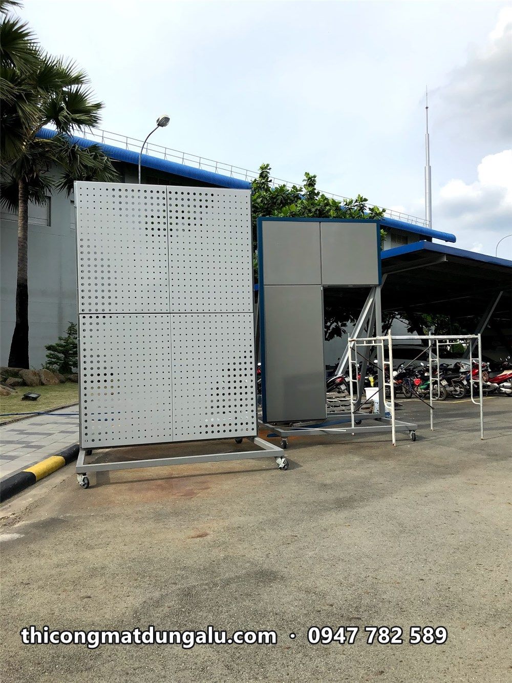 Tấm alu đục lỗ mẫu dựng trên chân sắt trong sân nhà máy, cạnh khối nhà xưởng