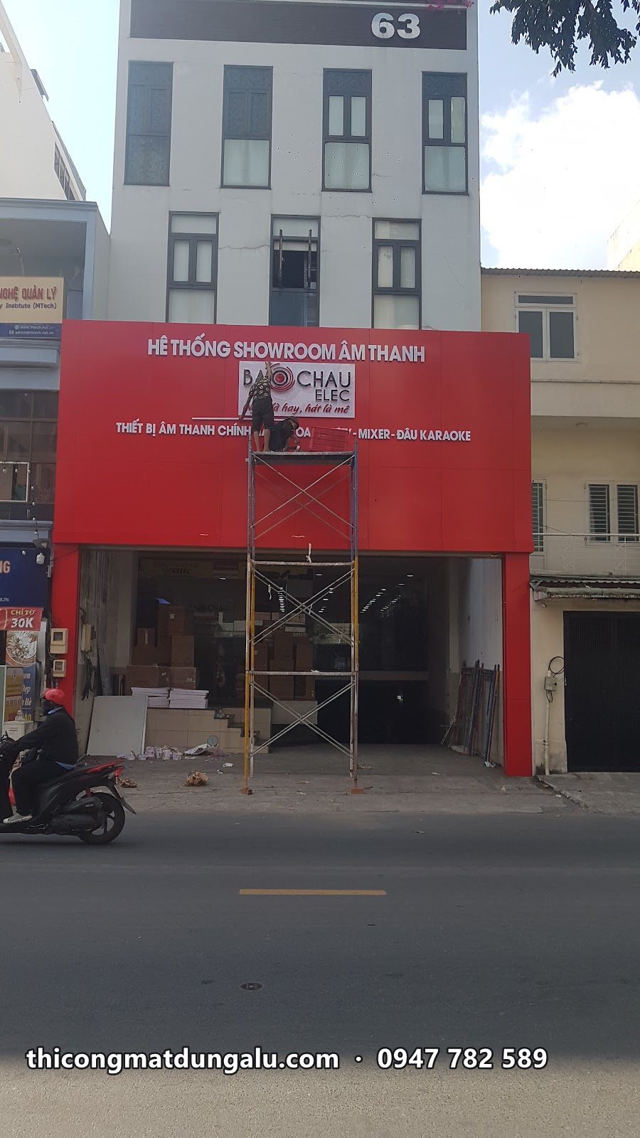 Tòa nhà nhiều tầng với mặt dựng đỏ tầng trệt đang lắp, giàn giáo phía trước