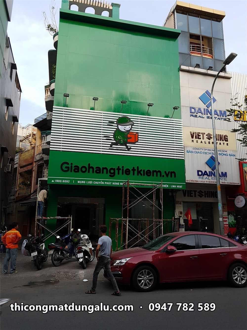 Bưu cục Giao Hàng Tiết Kiệm ba tầng ốp alu xanh kín, lam trắng và nhân vật ở tầng hai