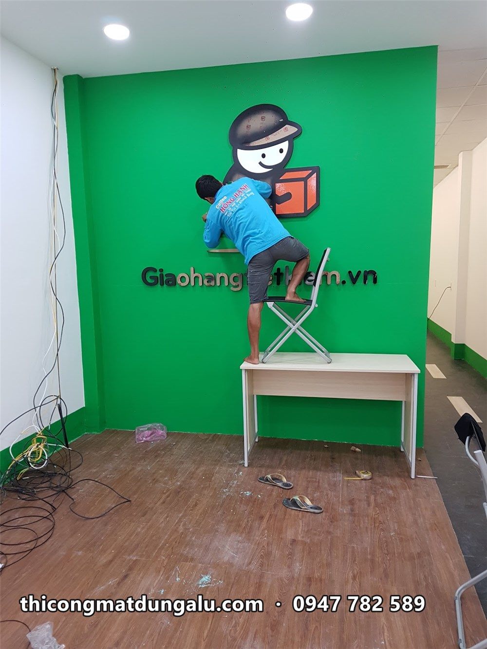 Thợ đứng trên bàn gắn nhân vật shipper lên tường xanh trong bưu cục