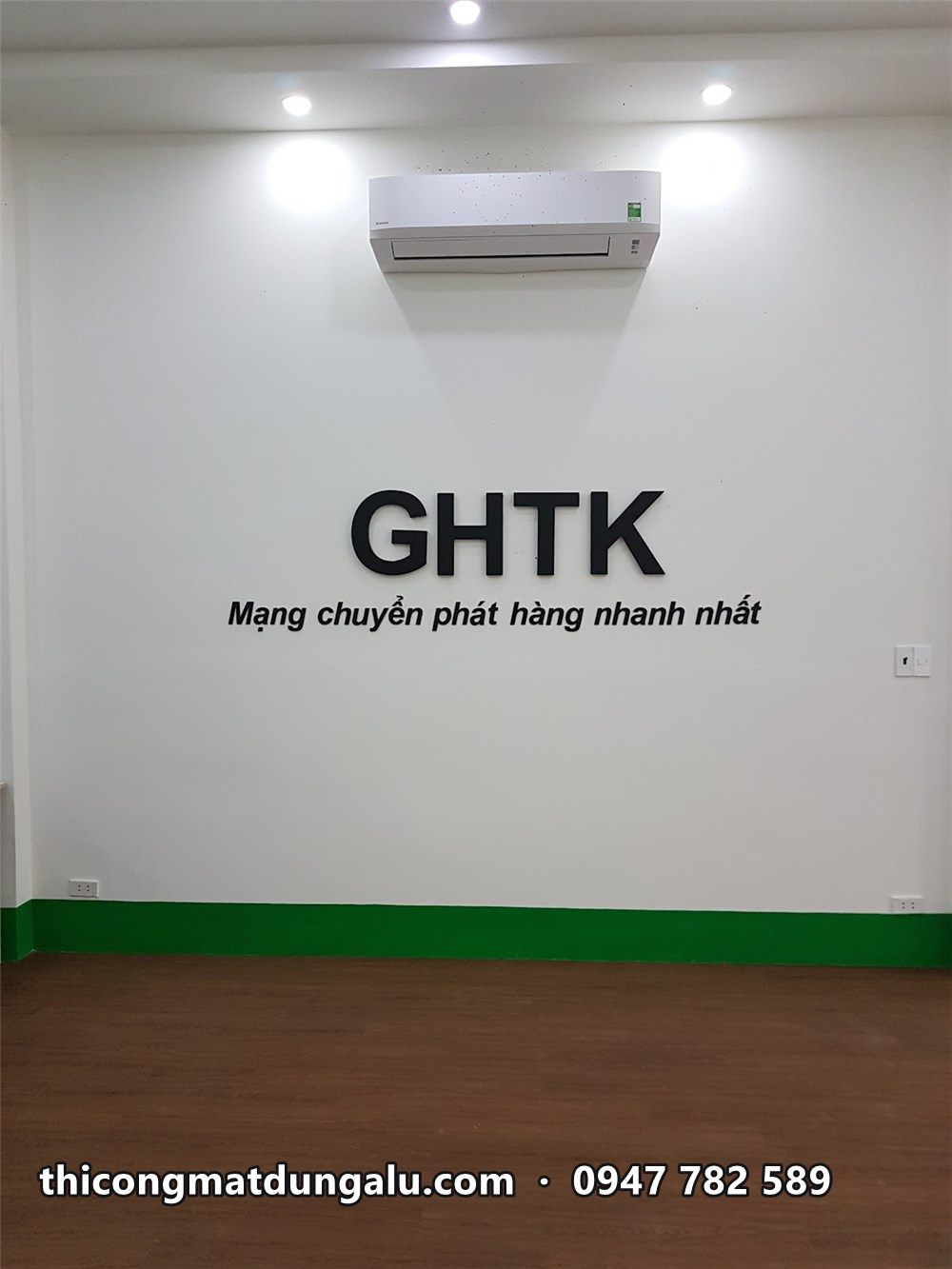 Chữ GHTK trên tường trắng phía trên máy lạnh, chân tường viền xanh