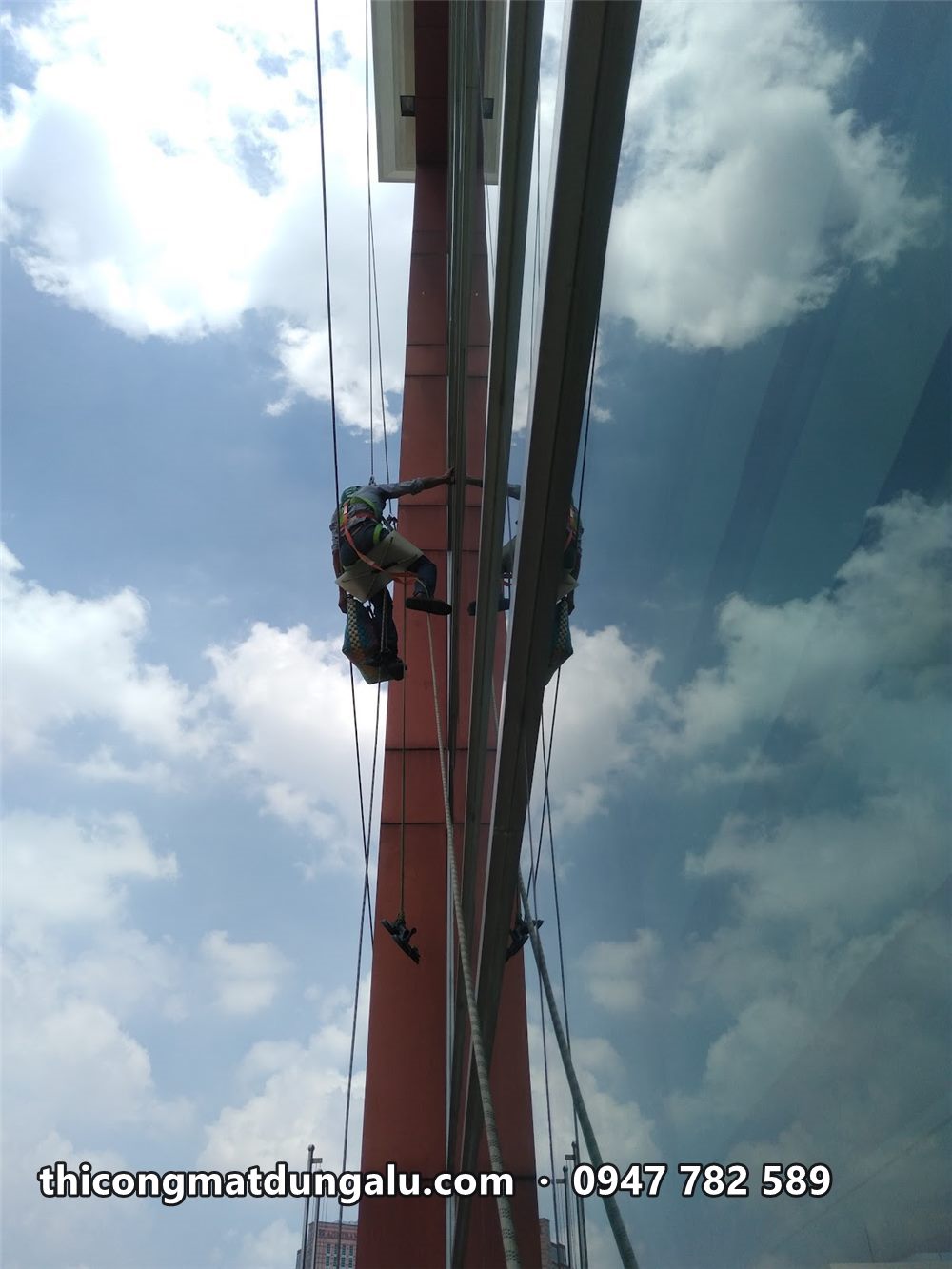 Thợ đu dây trên cao ốp alu màu nâu đỏ dọc cột tòa nhà, nền trời xanh mây trắng