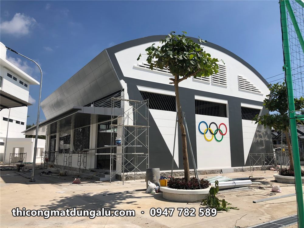 Nhà thi đấu mái vòm hoàn thiện với logo vòng tròn Olympic trên tường trắng và cây xanh phía trước