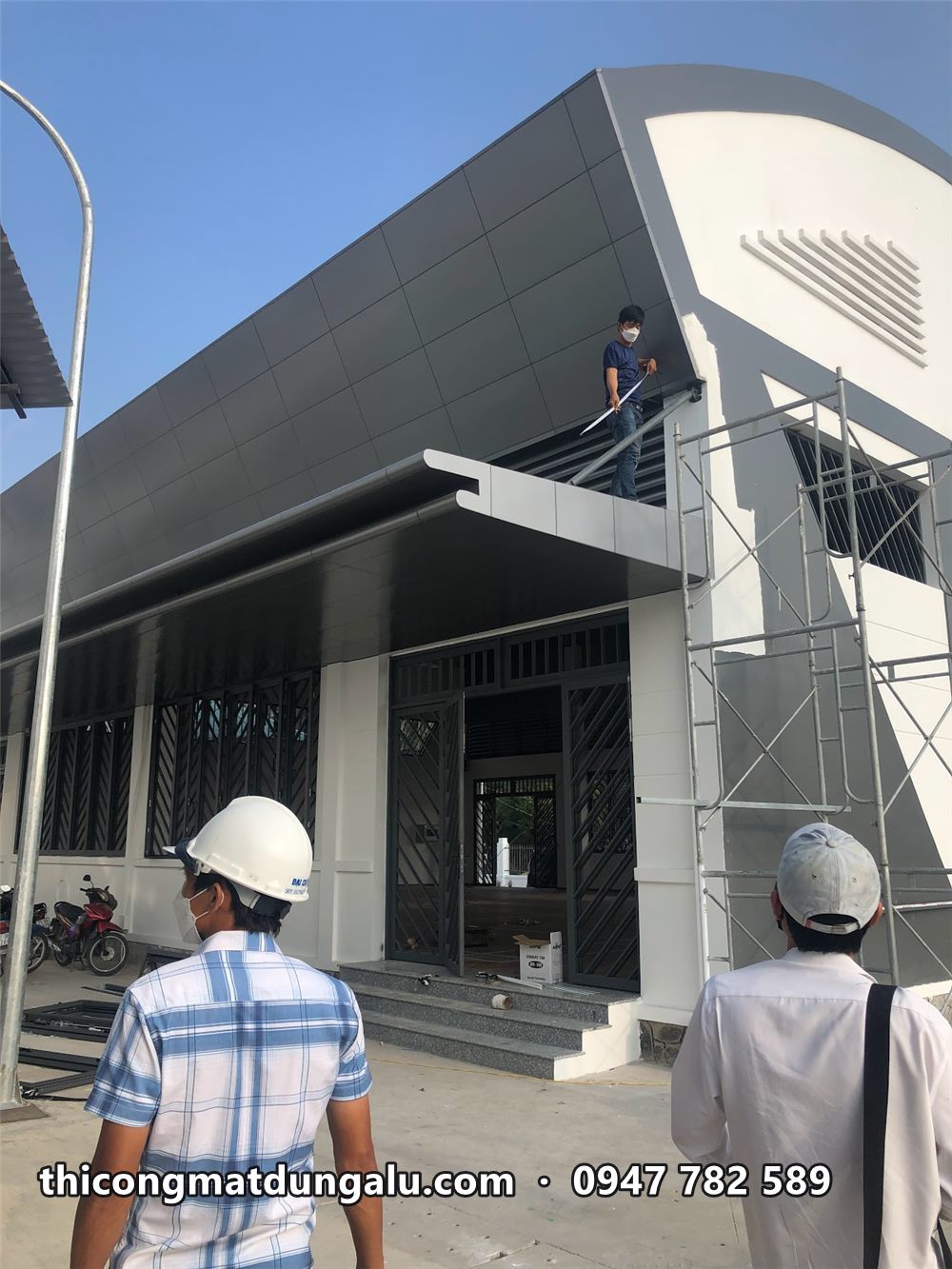 Lối vào nhà thi đấu với mái đón alu, hai kỹ sư đội nón bảo hộ trắng đứng nghiệm thu
