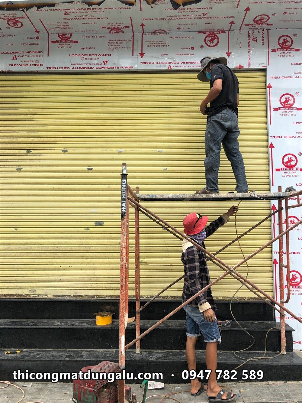 Thợ trên giàn giáo xử lý cửa cuốn vàng, tường đá hai bên phủ màng