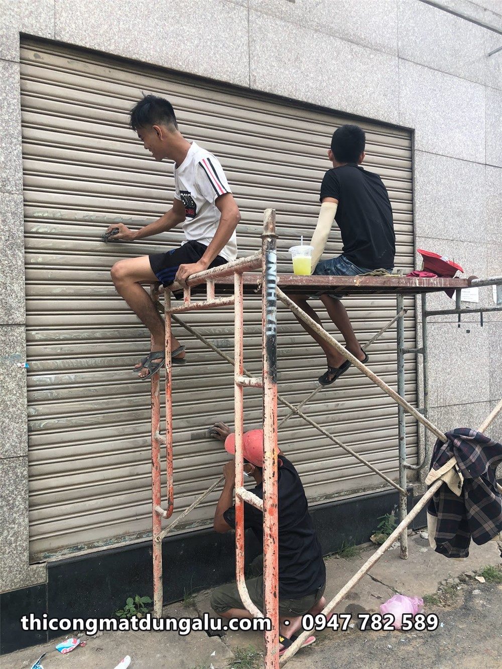 Hai thợ ngồi trên giàn giáo làm sạch cửa cuốn xám cũ