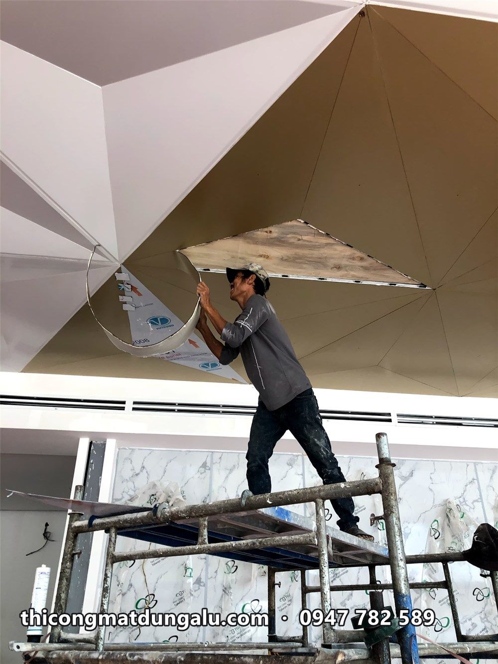 Thợ trên giàn giáo nâng tấm tam giác vàng đồng còn màng lên trần