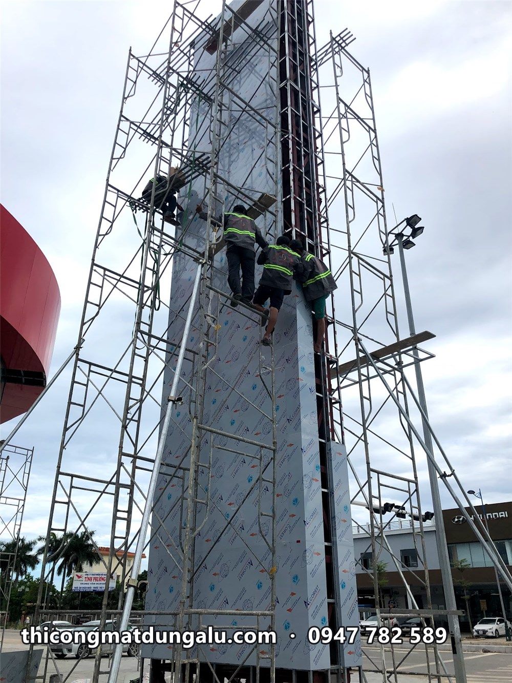 Thợ đứng trên giàn giáo bọc lớp tấm nền xám quanh trụ pylon